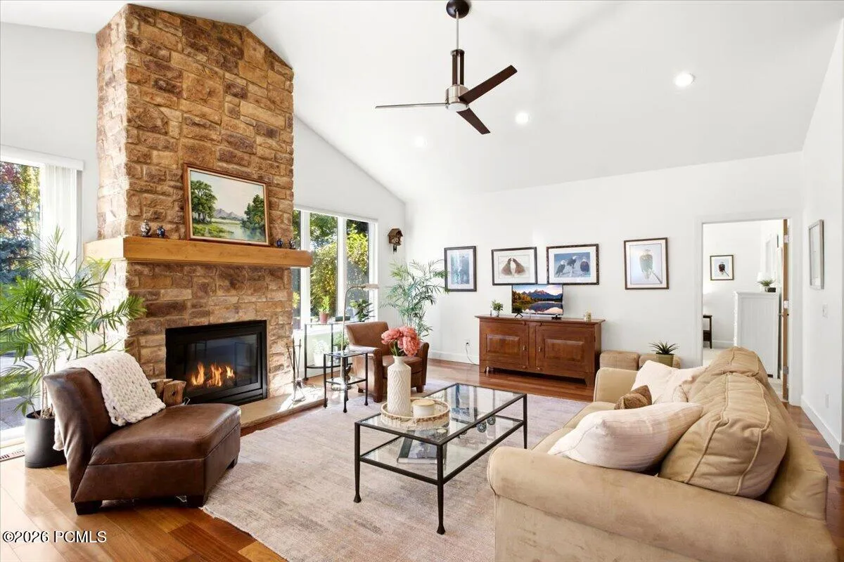 Living room featuring a Vaulted ceiling, stone fireplace with hearth, wood-finish flooring, and expansive windows