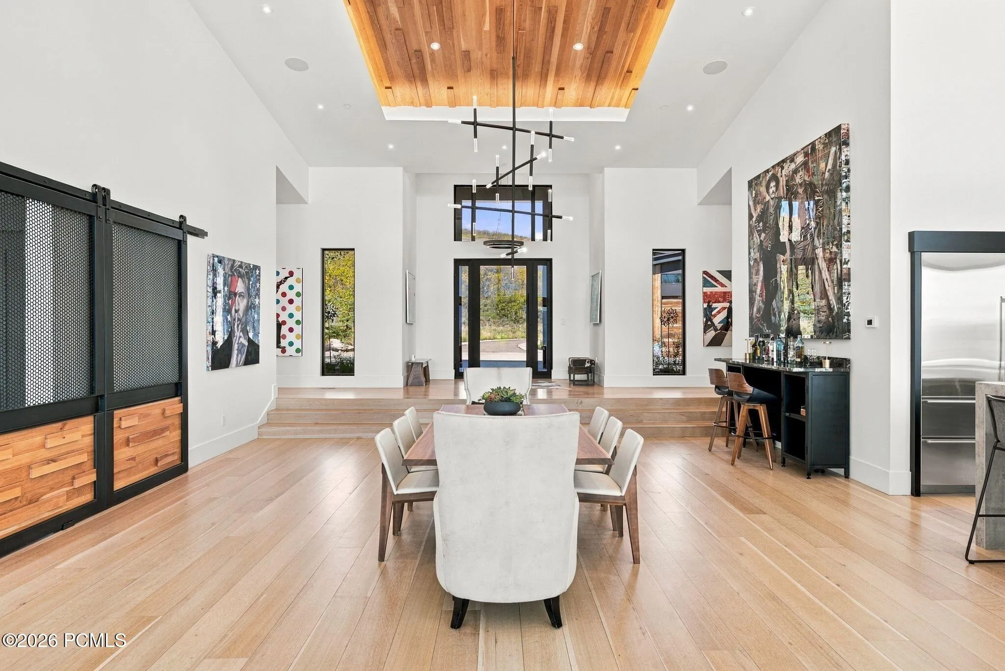 High-ceiling space featuring wood-finish flooring, a wood-paneled tray ceiling, custom black-framed sliding barn doors with wood inlay, and a multi-tiered modern pendant light fixture
