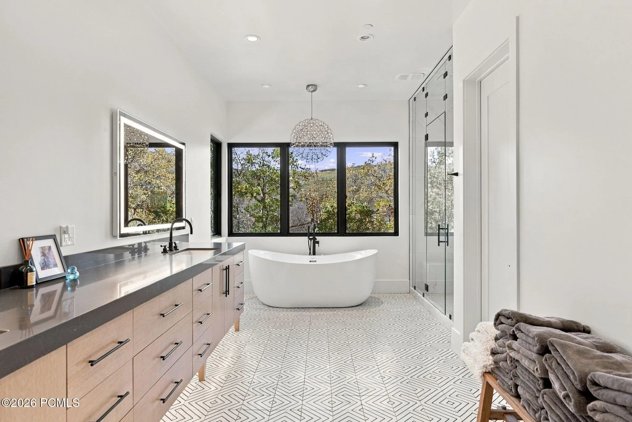 Primary bathroom featuring dual lighted vanities, freestanding soaking tub, glass-enclosed steam shower with full-height stone-finish wall tile, patterned geometric floor tile, and oversized windows to capture mountain views