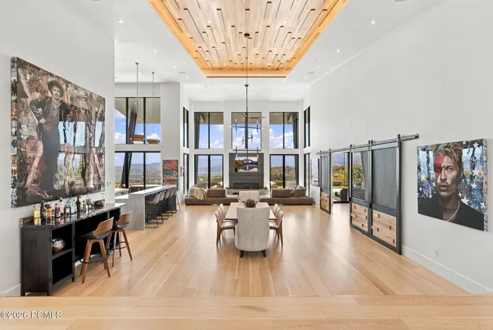 Expansive open-concept living area featuring custom wood-slat tray ceiling, floor-to-ceiling windows, light wood-finish flooring, bar, and sliding metal-framed barn doors