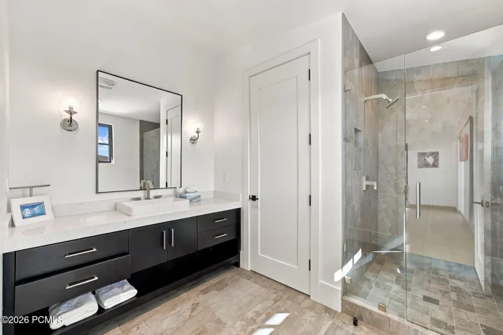 Modern vanity in office bathroom with dark cabinetry, vessel sink, and quartz countertop, paired with a walk-in shower featuring glass enclosure, recessed shelving, and stone-finish tile surround