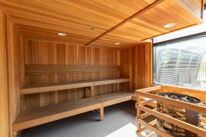 Built-in sauna featuring light wood paneling, multi-level wooden benches, a large exterior window, and a modern sauna heater with stone elements