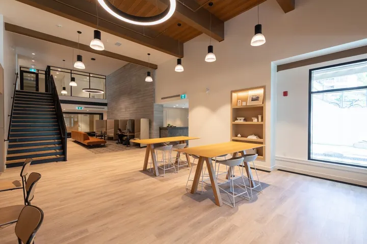 Spacious common area featuring light wood-finish flooring, exposed wood beam ceiling, and a prominent staircase with black metal railings