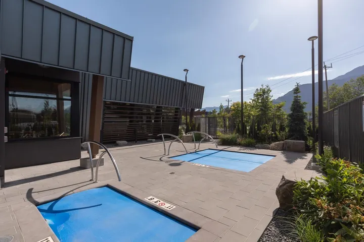 Outdoor amenity space featuring two covered pools, a paver patio, contemporary building architecture with dark paneling, and mature landscaping