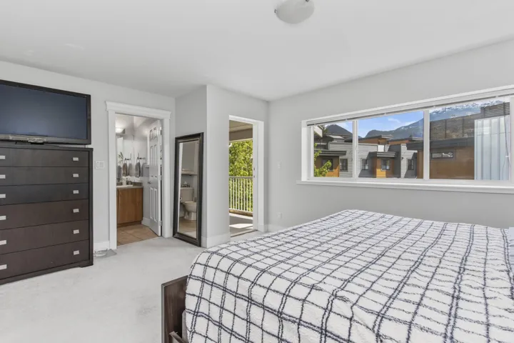 Carpeted bedroom with a large window, a private balcony, a ceiling-mounted light fixture, and an en-suite bathroom featuring a wood-finish vanity