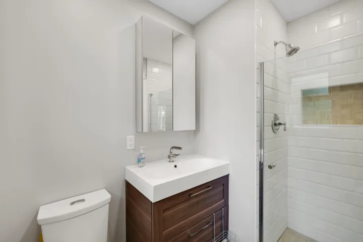 Contemporary bathroom featuring a wood-finish vanity with an integrated white sink, a mirrored medicine cabinet, a toilet, and a glass-enclosed shower with white subway tile