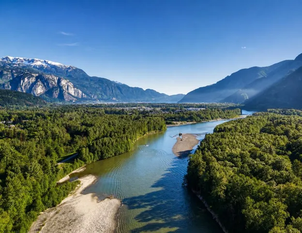 Expansive natural landscape featuring a broad river, dense evergreen forests, and distant snow-capped mountains