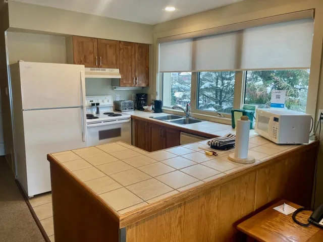Kitchen with brown cabinetry, white appliances, tile counters, under cabinet range hood, and a textured ceiling
