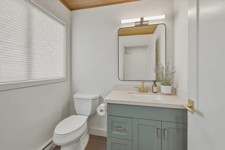 Powder room featuring a wood-paneled ceiling, a vanity with a light-colored countertop and a gold-finish faucet, a rectangular mirror with a metal frame, and a white toilet