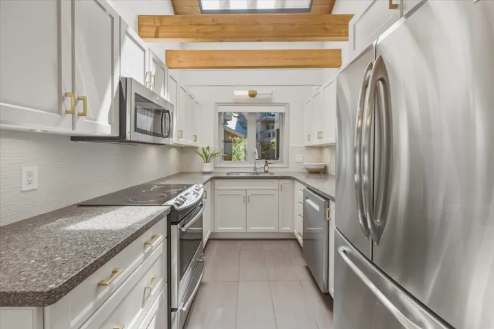 Modern galley kitchen featuring white cabinetry with gold-toned hardware, stainless steel appliances, dark speckled countertops, and a white subway tile backsplash