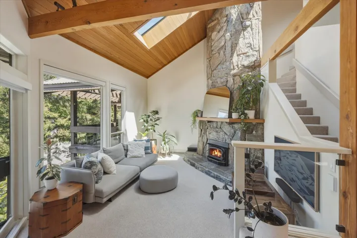 Vaulted wood-paneled ceiling with exposed beams and skylight, stone-clad fireplace with wood stove, large glass sliding doors, carpeted flooring, and an open staircase with glass panel railing