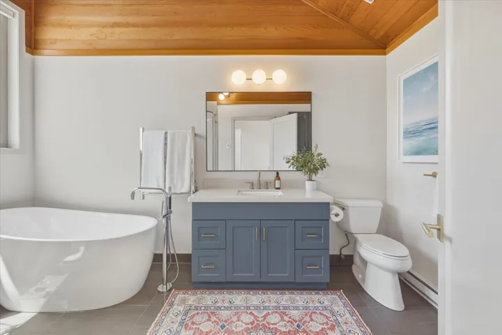 Freestanding soaking tub with floor-mounted faucet, wood-paneled vaulted ceiling, blue vanity with white countertop, wall-mounted mirror, and tile flooring