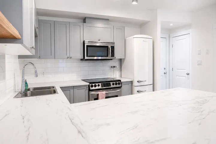 Kitchen featuring light grey cabinetry, white subway tile backsplash, stainless steel appliances, a double basin sink with a gooseneck faucet, and white marble-look countertops