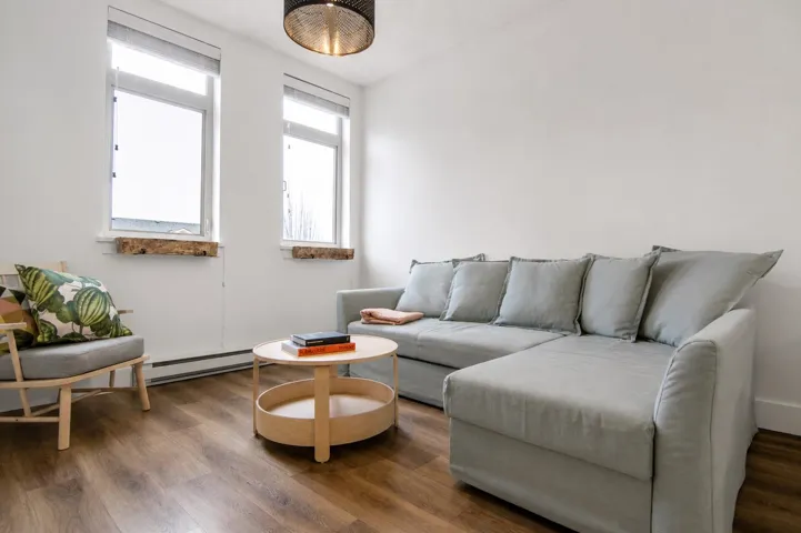 Bright living area featuring wood-finish flooring, white walls, and two large windows with roller shades
