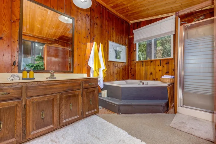Wood-paneled bathroom featuring a built-in vanity with cabinetry, a drop-in bathtub with tiered surround, a glass-enclosed shower, and a window with blinds