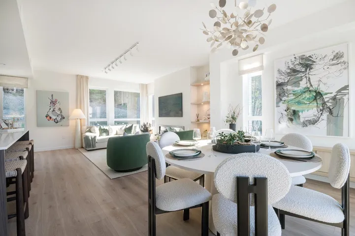 Open-concept living and dining area featuring light wood-finish flooring, a contemporary chandelier, large windows with natural light, a built-in shelving unit with recessed lighting, and a kitchen island with seating