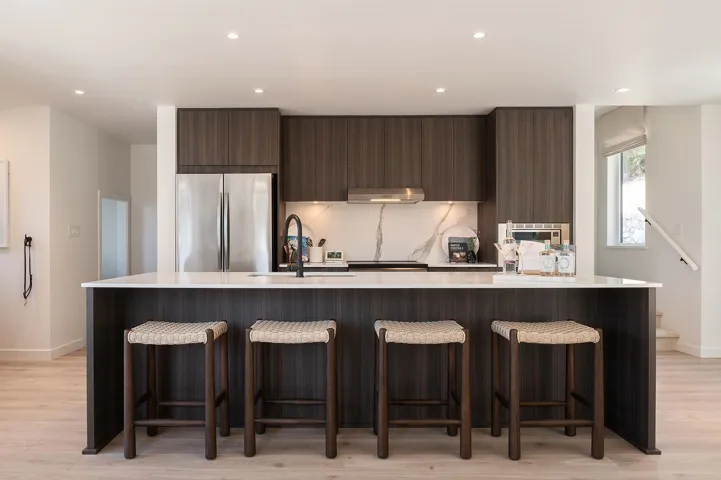 Modern kitchen featuring a large island with white countertop, dark wood-finish cabinetry, stainless steel appliances, and a marble-patterned backsplash