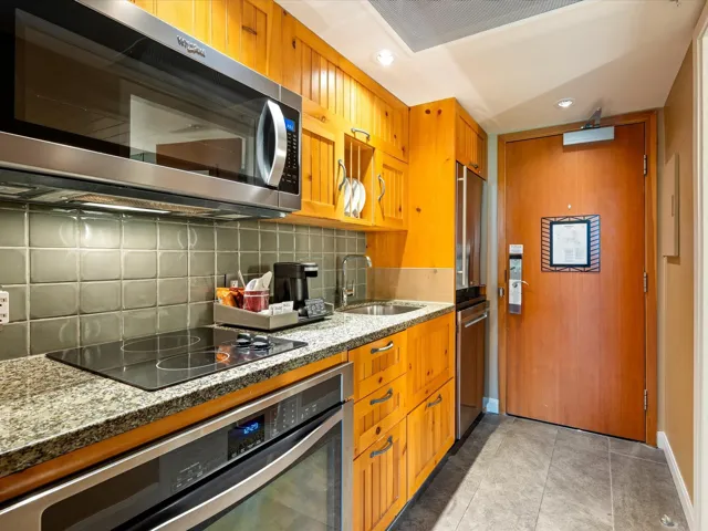 Kitchen featuring stainless steel appliances, backsplash, brown cabinetry, stone countertops, and light tile patterned floors