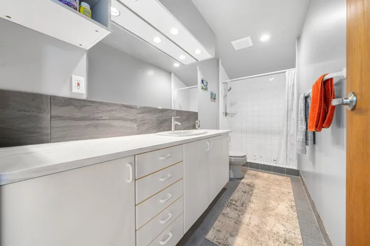 Spacious bathroom featuring a white vanity with integrated sink, extensive countertop space, recessed lighting, a tiled shower enclosure, and gray floor tiling