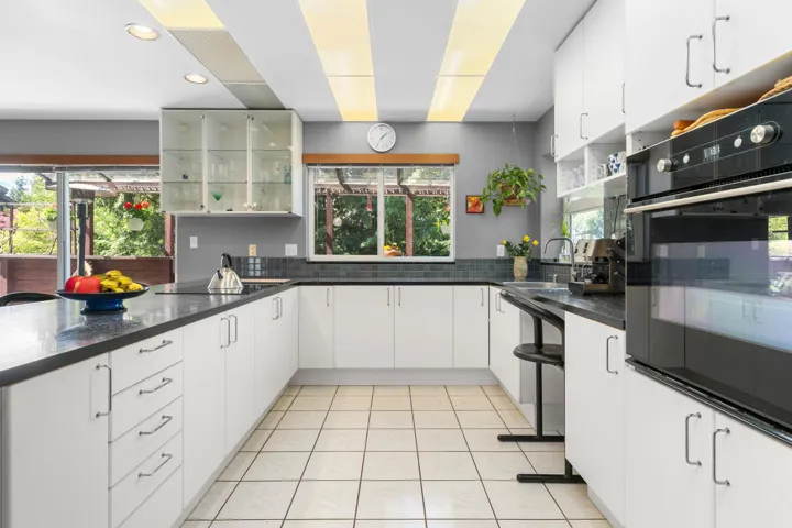 Bright kitchen featuring white cabinetry, dark countertops, tiled flooring, built-in oven, and a glass-front display cabinet