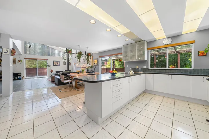 Open-concept kitchen and living area featuring white tile flooring, a large island with black countertop, white cabinetry with glass-front uppers, and a window above the sink