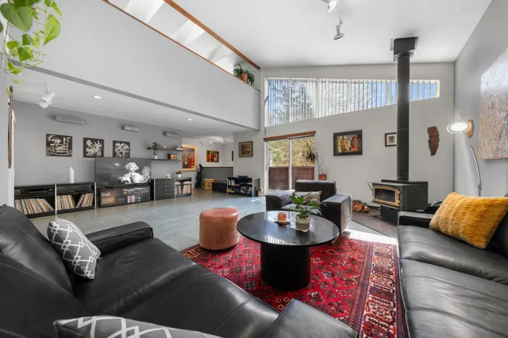 Spacious living area featuring a vaulted ceiling with skylights, an upper-level architectural opening, a dark wood-burning stove, large sliding glass doors, and light gray tile flooring