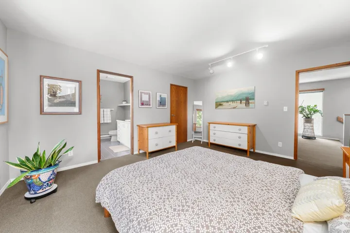 Carpeted bedroom featuring light gray walls, track lighting, and a doorway opening to a bathroom with tile flooring and a white vanity