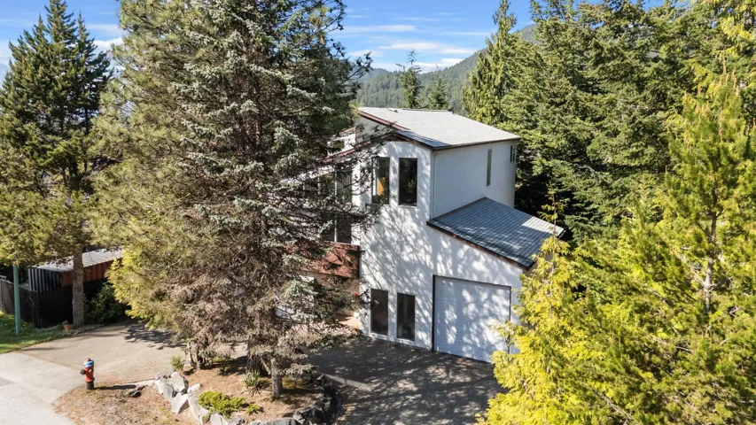 Contemporary architecture with a white stucco facade, a two-car garage, multiple large windows, and a surrounding natural landscape with mature evergreen trees and distant mountains
