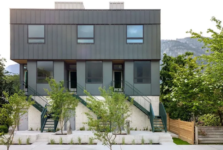 Contemporary multi-story exterior featuring dark gray paneling, large windows, and green metal staircases leading to individual entrances