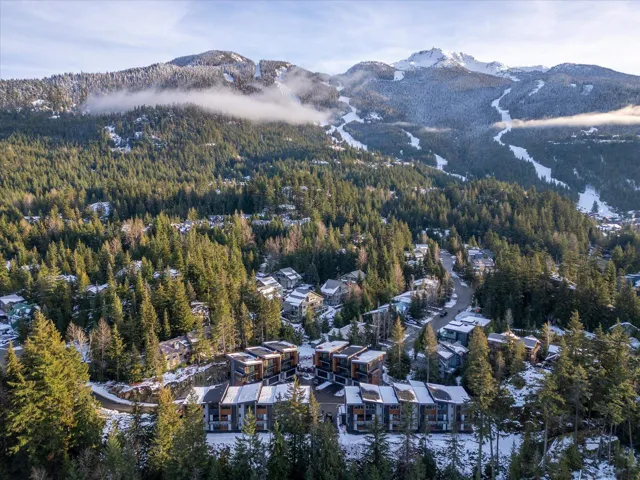 Contemporary residential buildings set amidst a coniferous forest, featuring snow-dusted ground and surrounding mountain peaks