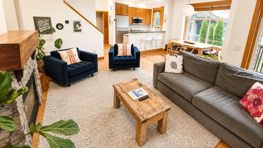Open-concept living area featuring wood-finish flooring, a stone-surround fireplace with a substantial wood mantel, a staircase with wood treads, a kitchen with wood cabinetry and stainless steel appliances, and a dining space with large windows