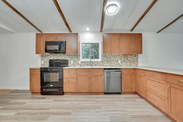 Kitchen with backsplash, black appliances, brown cabinetry, light wood finished floors, and beam ceiling