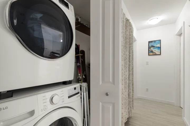 Stackable front-load washer and dryer set, complemented by a hallway featuring light-toned wood-finish flooring and a flush-mount ceiling light fixture