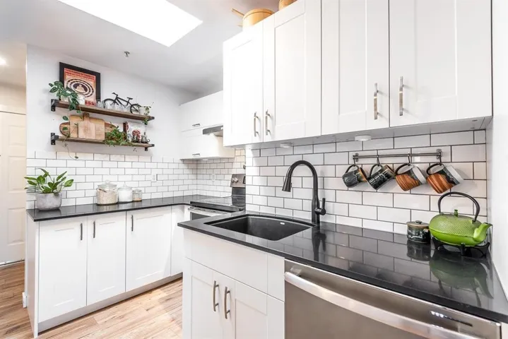 Modern kitchen featuring white shaker cabinetry, dark countertops, white subway tile backsplash, a black sink with a matching faucet, and wood-finish flooring