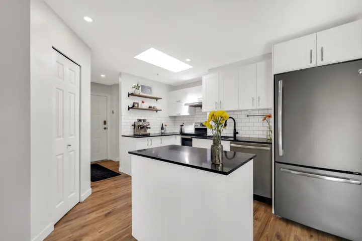 Modern kitchen featuring a central island with a dark countertop, stainless steel appliances, white cabinetry, wood-finish flooring, and a skylight