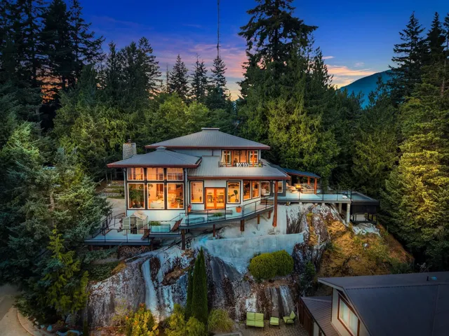 Contemporary residence featuring a metal roof, extensive window walls, multiple outdoor decks with glass railings, and a prominent stone chimney, set amidst dense evergreen trees