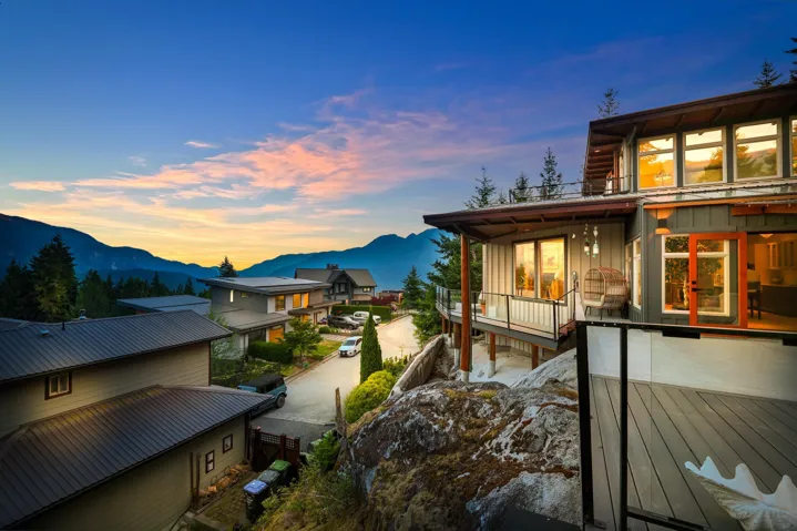 Contemporary multi-level residence featuring an expansive outdoor deck, a balcony with glass railings, large windows, and a multi-pitched roofline, all set against a mountainous backdrop