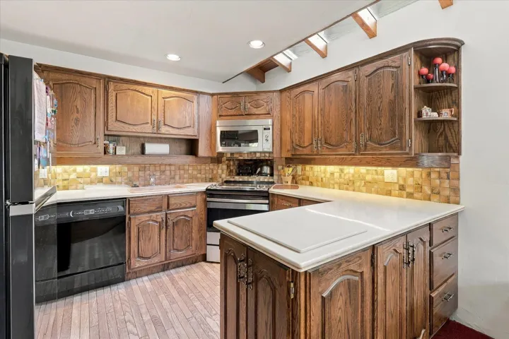 Wood-finish cabinetry, light-toned countertops, tile backsplash, built-in microwave, and wood-finish flooring