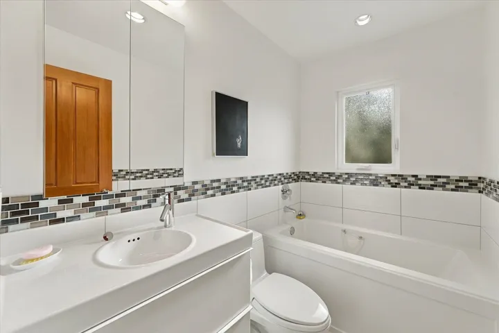 White vanity with integrated sink, mosaic tile backsplash, recessed lighting, white tiled tub, and a window with privacy glass