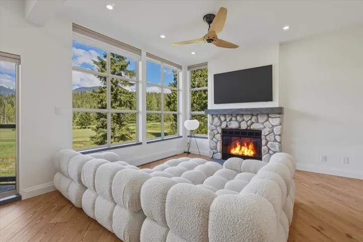 Living area with a stone fireplace, wood-finish flooring, a wood-blade ceiling fan, and large picture windows