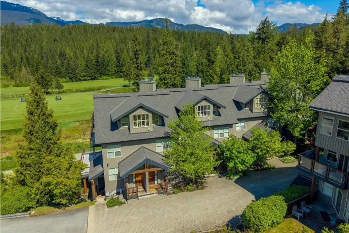 Multi-level residential structure with multiple dormer windows, a covered entry, and a pitched roof, situated adjacent to a green space, with surrounding mature trees and distant mountains