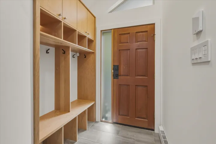 Wood-finish entry door with frosted sidelight, built-in wooden storage unit with bench seating, overhead cabinetry, and wall-mounted coat hooks, all set against neutral wall tones and tile flooring
