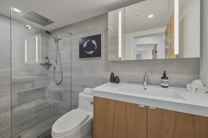 Contemporary bathroom featuring a glass-enclosed shower with tiled walls and built-in shelving, a wood-finish vanity with a white countertop and integrated sink, and an illuminated mirrored medicine cabinet