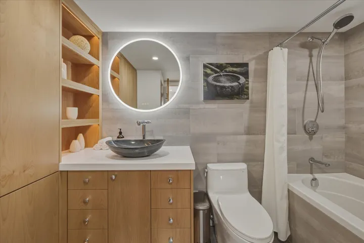 Bathroom featuring wood-finish cabinetry, a round illuminated mirror, a vessel sink, a white countertop, and a tiled shower/tub combination
