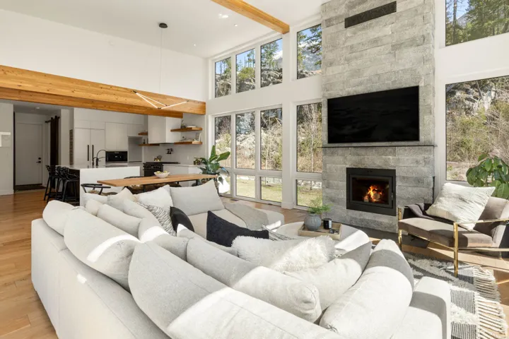 Open-concept living space featuring a floor-to-ceiling stone fireplace, expansive windows, wood-finish flooring, exposed wood ceiling beams, and a modern kitchen with a large island and open shelving