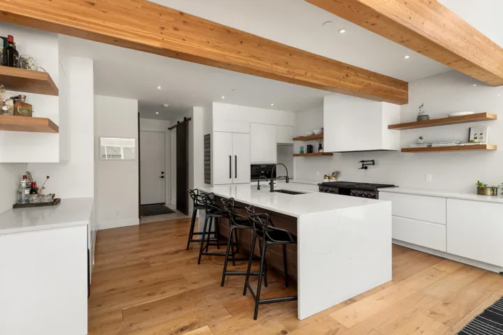 Modern kitchen featuring a large waterfall-edge island with seating, exposed wood ceiling beams, wood-finish flooring, white flat-panel cabinetry, and open shelving