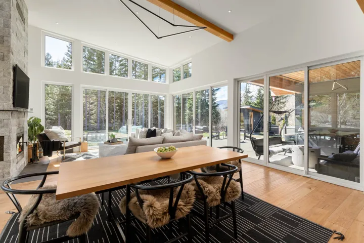 Open-concept living space featuring a floor-to-ceiling stone fireplace, expansive window walls, light wood-finish flooring, and exposed ceiling beams