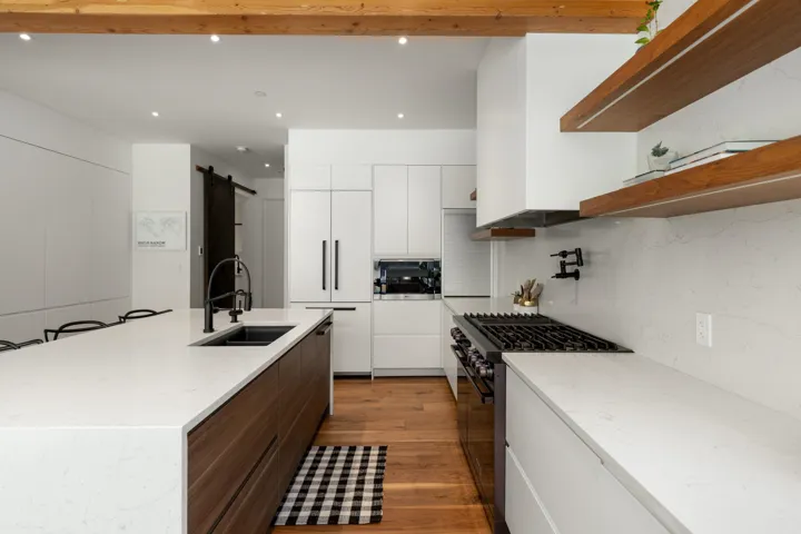 Modern kitchen featuring a large island with an integrated sink and matte black faucet, wood-finish flooring, white cabinetry, integrated appliances, and exposed wood beams