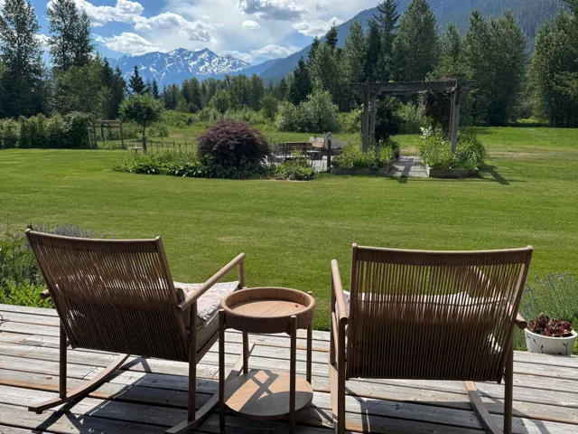 Expansive outdoor decking features a broad lawn with a garden bed and a wooden pergola, all framed by distant mountains and surrounding evergreen trees