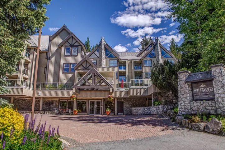 Multi-story building featuring stone and wood siding, multiple balconies with glass railings, and a prominent peaked roofline